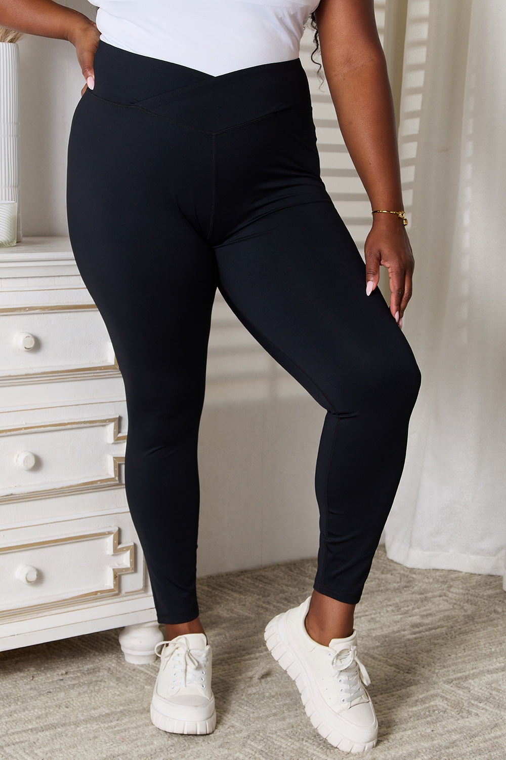 A person is standing in a room wearing Basic Bae V-Waistband Sports Leggings that offer a flattering fit, paired with white slip-on shoes. To the left, there is a white dresser with drawers. This outfit highlights an active lifestyle, seamlessly blending comfort and style.