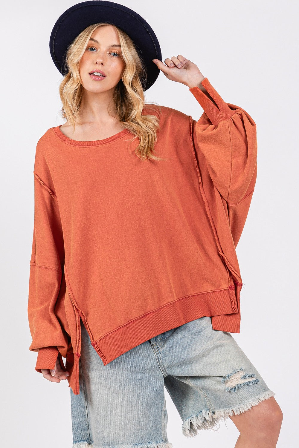 A person wearing a wide-brimmed hat, the SAGE + FIG Mineral Wash Side Slit Oversized Sweatshirt, and frayed denim shorts stands against a plain background.