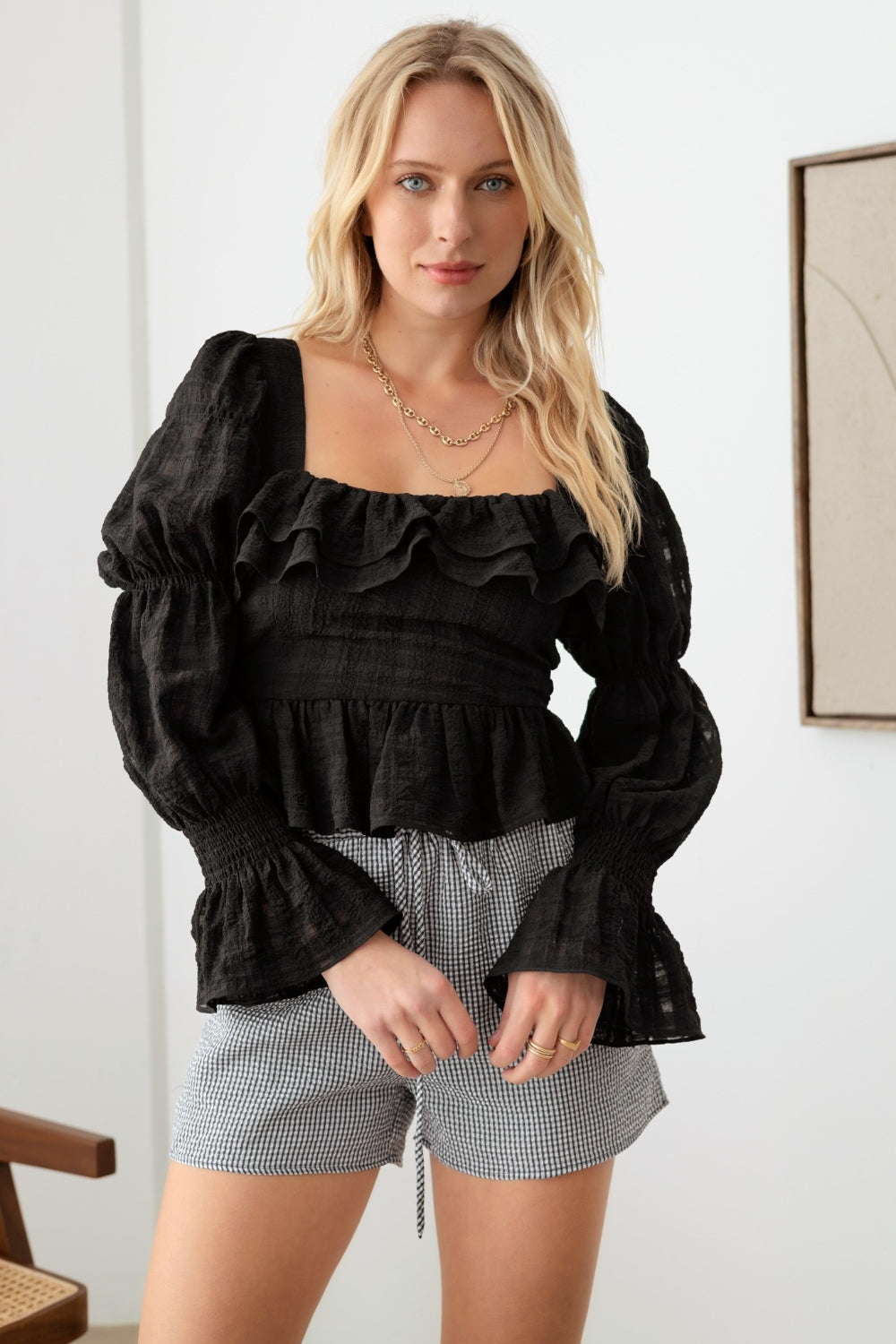 A person with long blonde hair is standing indoors, wearing a Mustard Seed Shirred Smocked Peplum Long Sleeve Top and checkered shorts for a trendy feminine look. They are gazing directly at the camera with hands resting lightly on their hips.