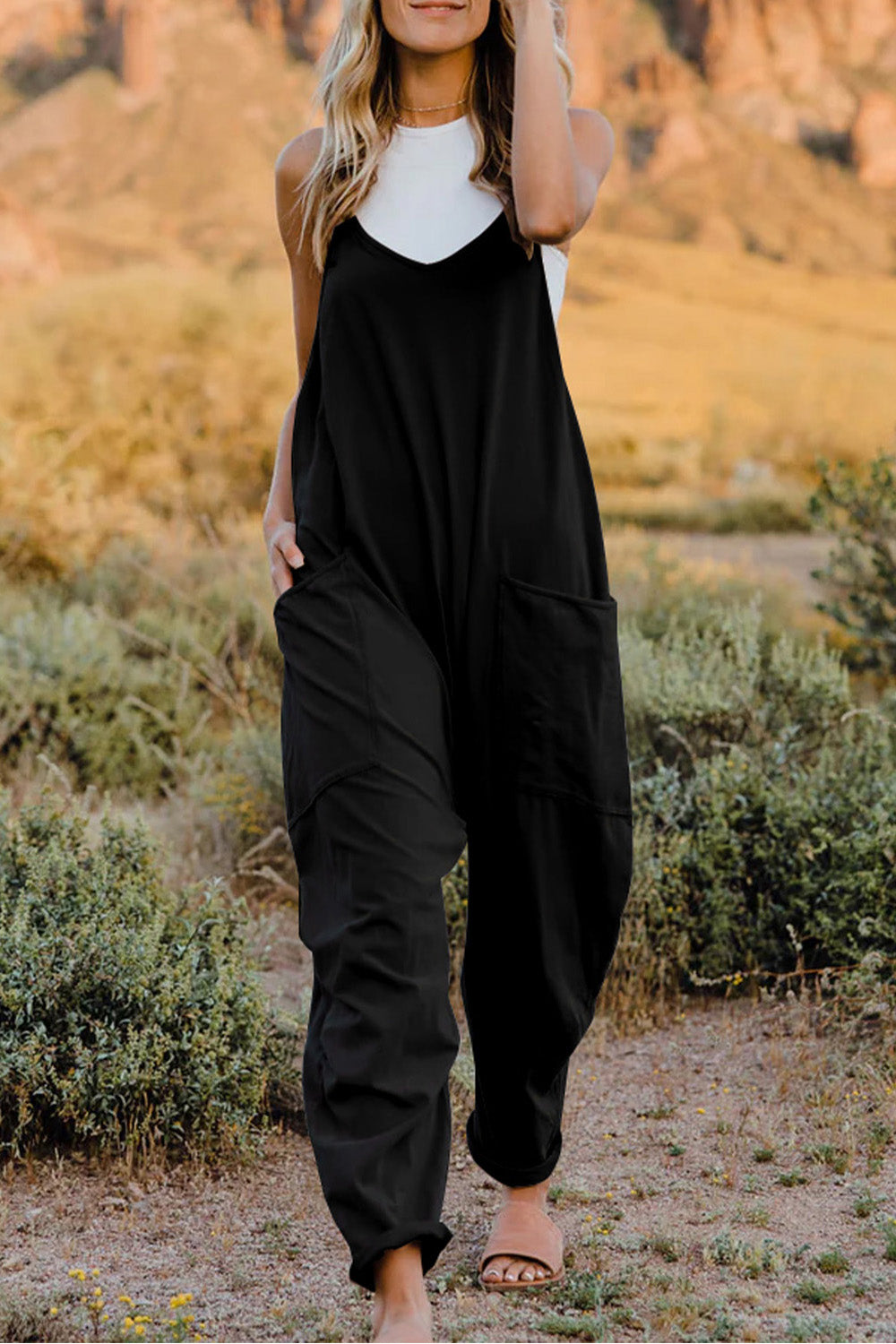 Person wearing a Double Take Full Size V-Neck Sleeveless Jumpsuit with Pockets and sandals, walking outdoors on a dirt trail with bushes in the background.