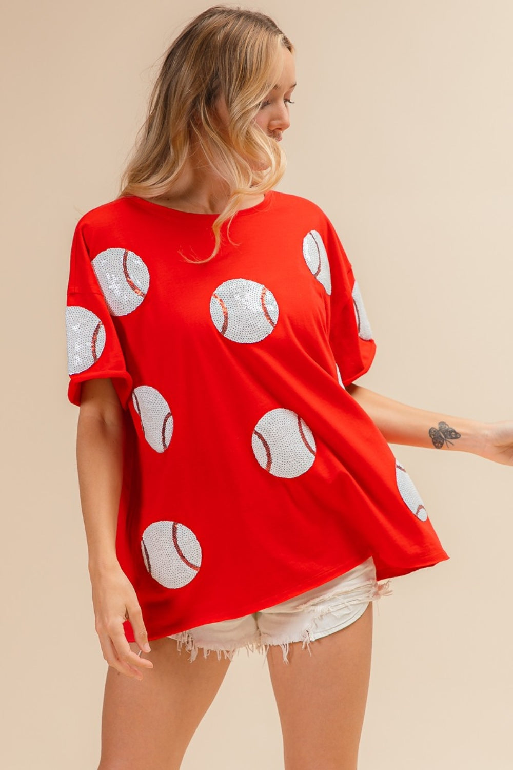 Woman wearing the BiBi Sequin Baseball Drop Shoulder Short Sleeve T-Shirt in red, paired with white shorts, posing against a beige background.