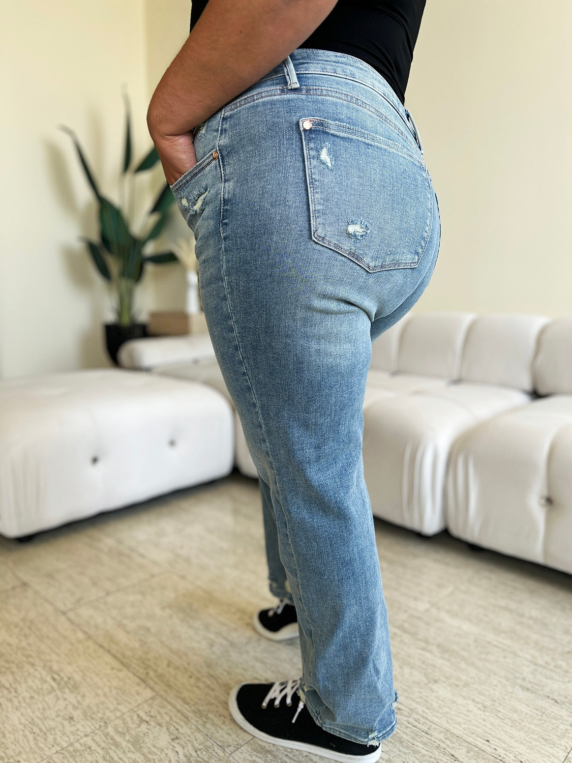 A person wearing Judy Blue Full Size Mid Rise Distressed Straight Jeans in light blue with a white top and white slip-on shoes stands in a room with a white couch and a plant.