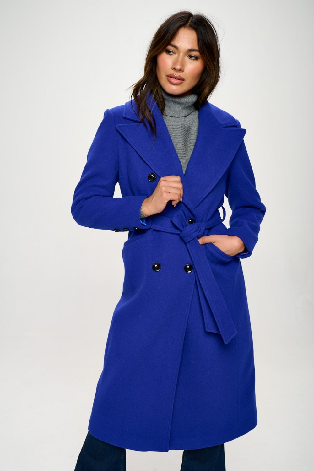 Person wearing the Coalition LA Double-Breasted Longline Coat with Belt, made from vegan wool in blue, over a grey turtleneck, posing against a plain background.