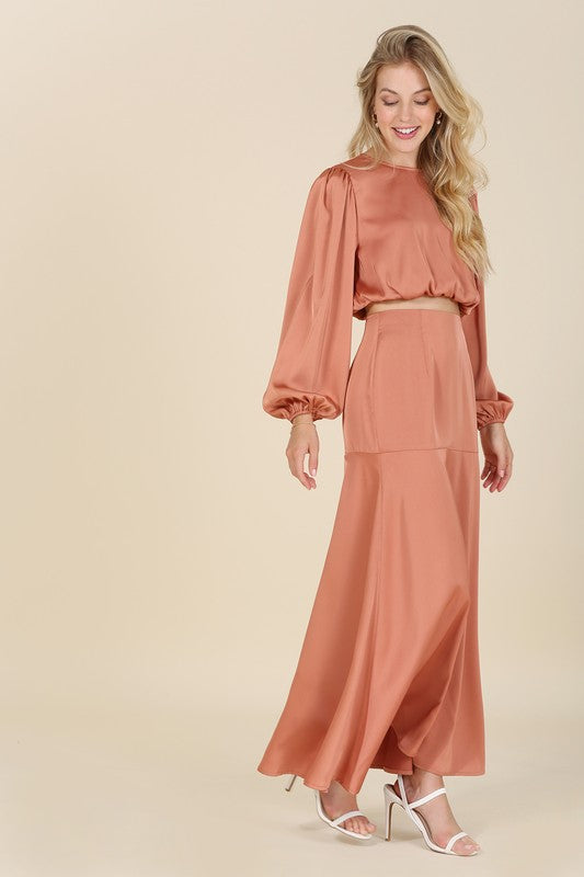 A woman stands against a plain background, elegantly adorned in the "Dressed up satin two-piece mermaid dress set" featuring a peach-colored, balloon-sleeve crop top and matching high-waisted maxi skirt, accessorized with white high-heeled sandals.