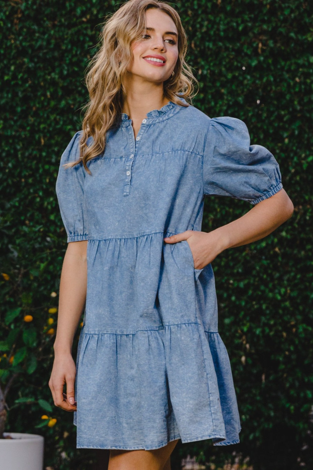 A person wearing the ODDI Washed Tiered Mini Denim Dress with puffed sleeves stands in front of a green leafy background.