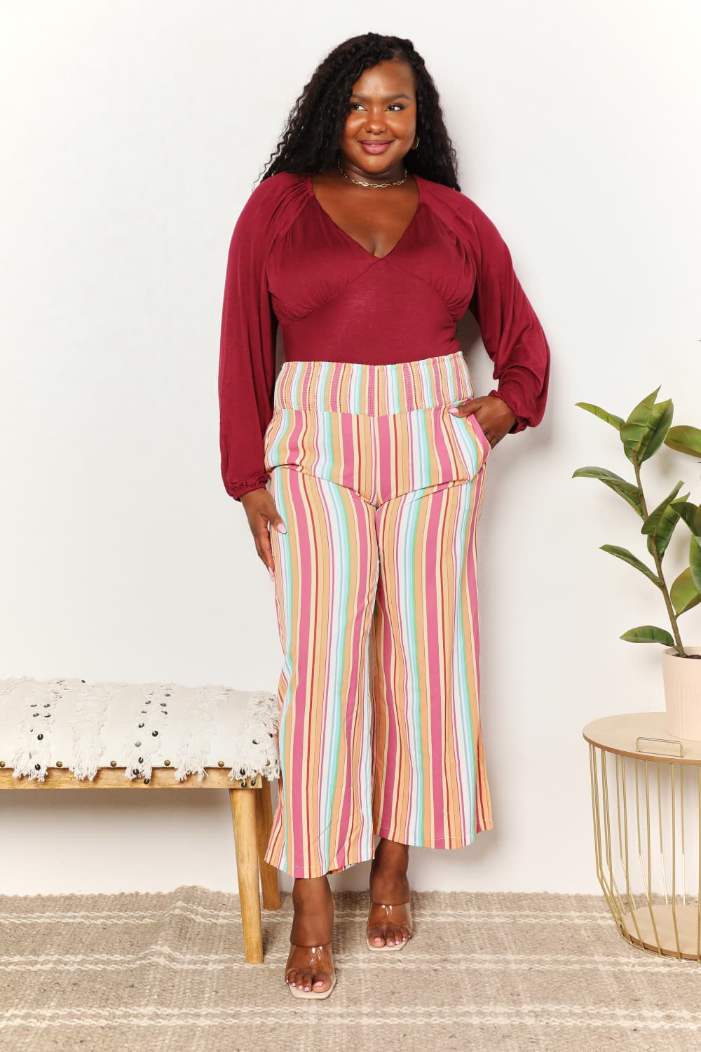 A person wearing Double Take Striped Smocked Waist Pants with Pockets and a burgundy long-sleeve shirt stands beside a wooden stool with a hat on it.