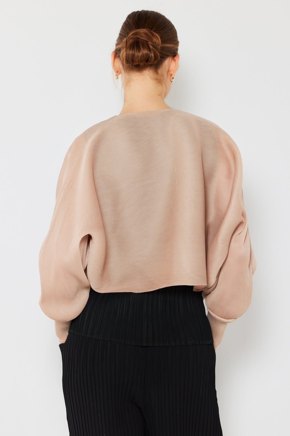 A person stands wearing the stylish Marina West Swim Rib Pleated Puff Sleeve Bolero Cardigan in beige, featuring draped sleeves, paired with a trendy black pleated top.