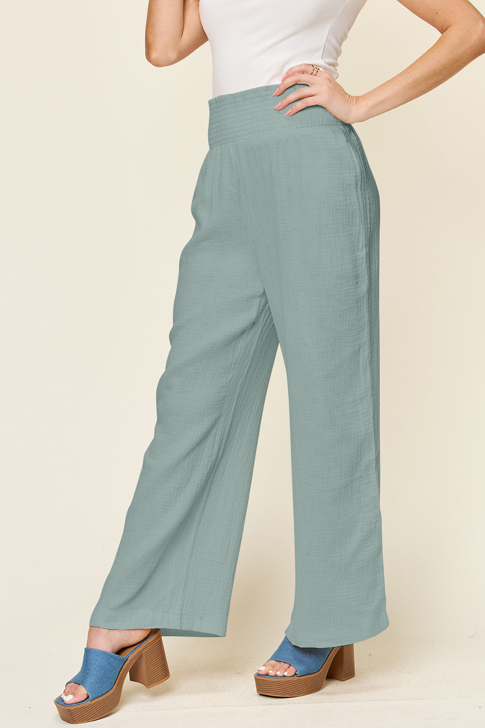 A person wearing Double Take Full Size Texture Smocked Waist Wide Leg Pants in black and a white 100% cotton top is standing with one hand on their hip. They are also wearing blue open-toe sandals.