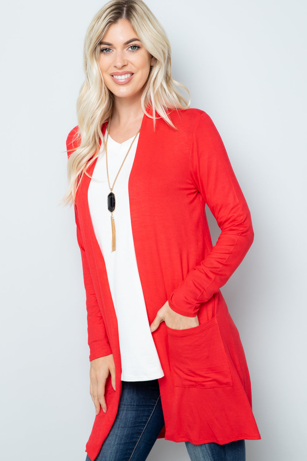 A woman in a highly stretchy Celeste Full Size Open Front Cardigan with Pockets, along with a white top and blue jeans, stands against a plain background, with one hand in her cardigan pocket, wearing a long necklace.