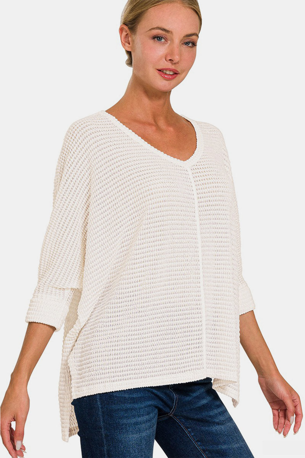 A smiling woman confidently poses in the Zenana Full Size Round Neck High-Low Slit Knit Top, which features a loose fit and three-quarter sleeves. She pairs the white knit top with blue jeans, striking a pose with one hand on her head and the other in her pocket.