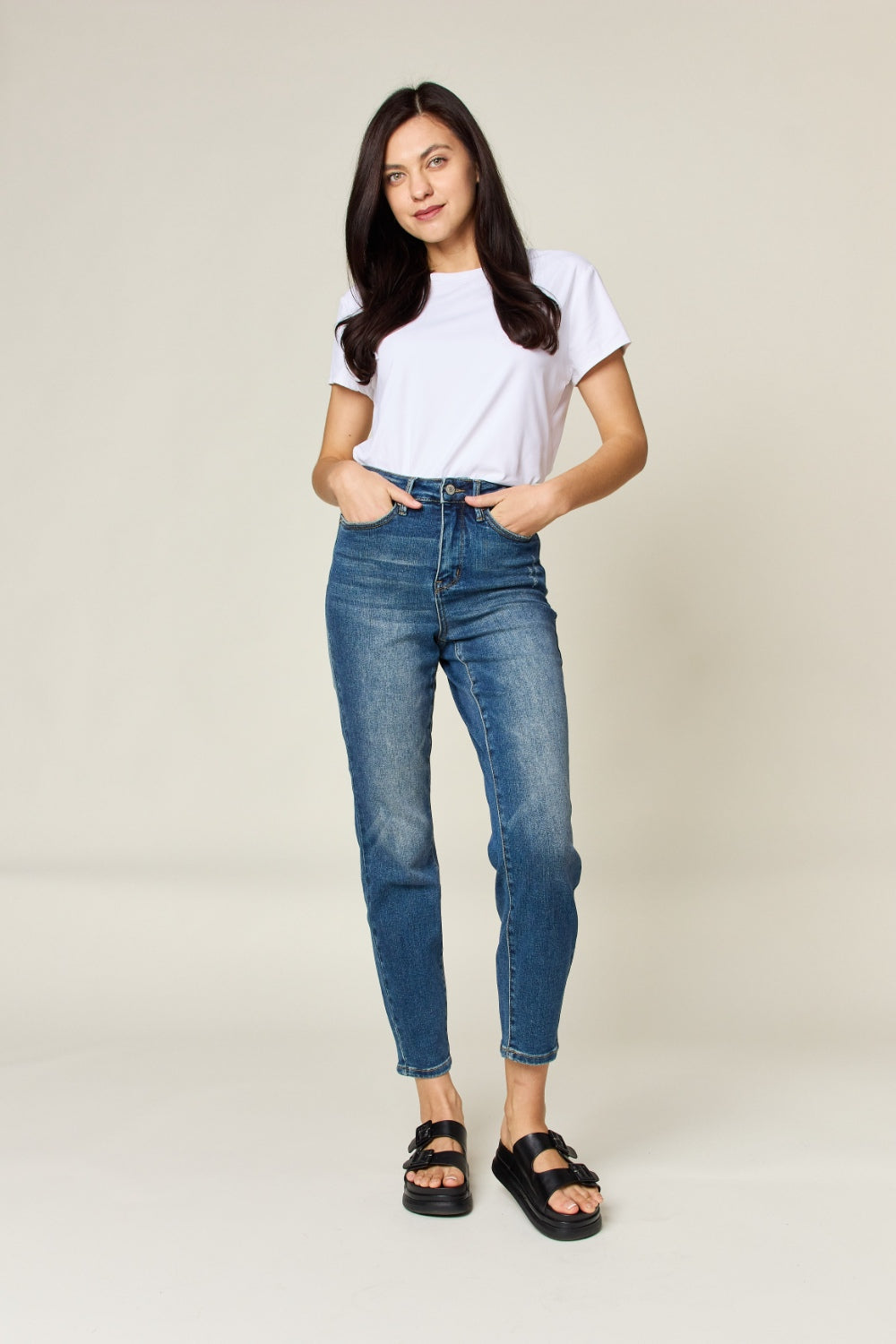 Person wearing Judy Blue Full Size Tummy Control High Waist Slim Jeans in blue and a white shirt, with their hands in their pockets. They are standing indoors on a polished concrete floor with a wooden cabinet and plant in the background.