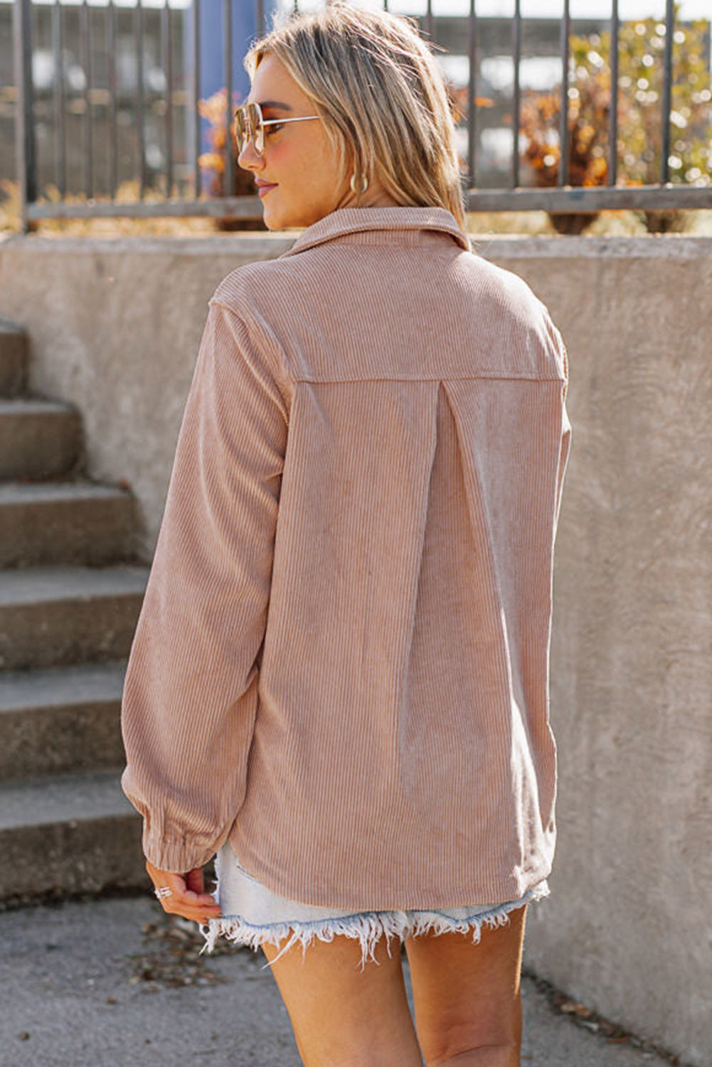 A person with long blonde hair, wearing a trendy Apricot Ribbed Texture Half Zip Collared Sweatshirt and blue jeans, stands facing away and looks over a stone balustrade.