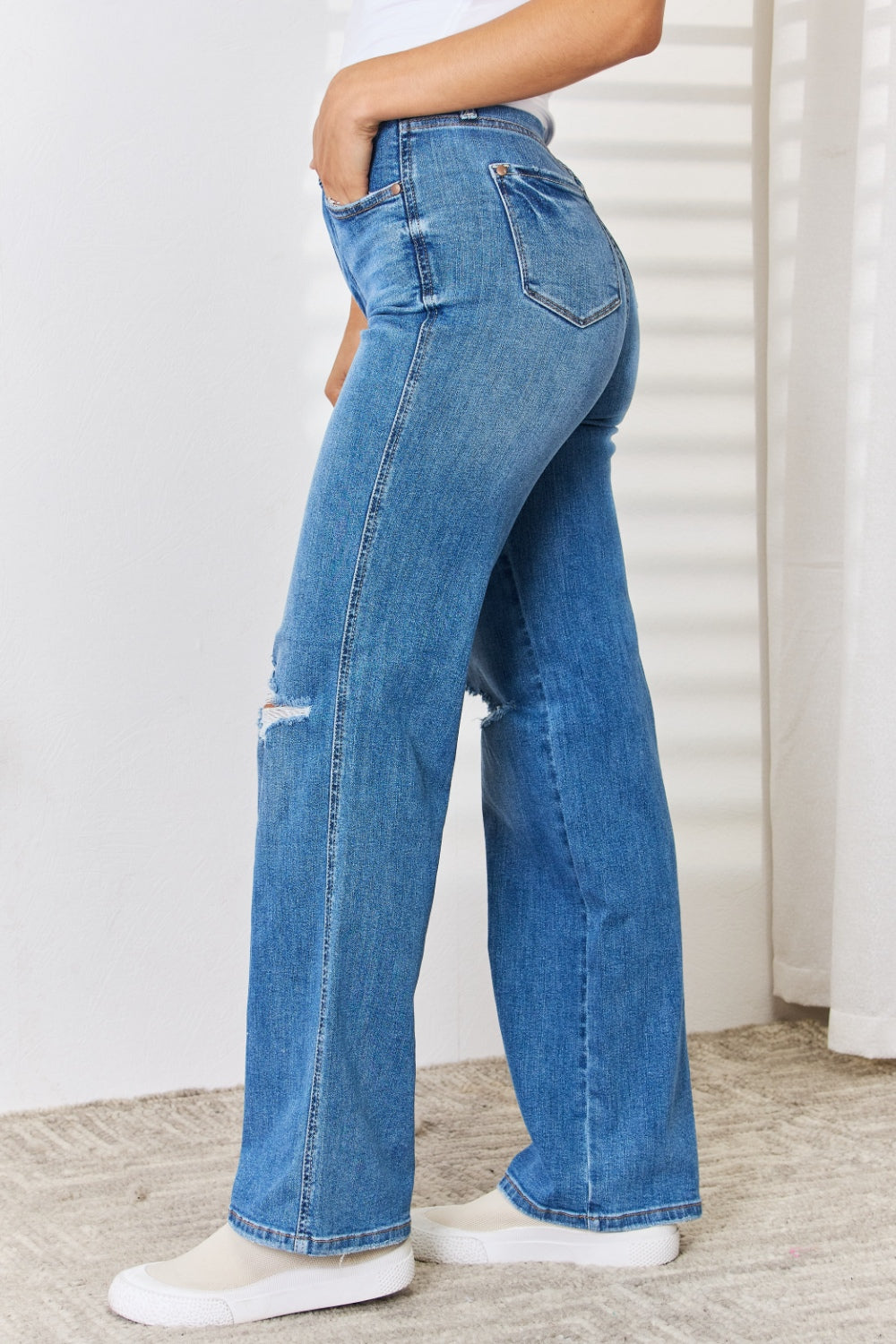 A person wearing the Judy Blue Full Size High Waist Distressed Straight-Leg Jeans and white sneakers stands against a white wall.