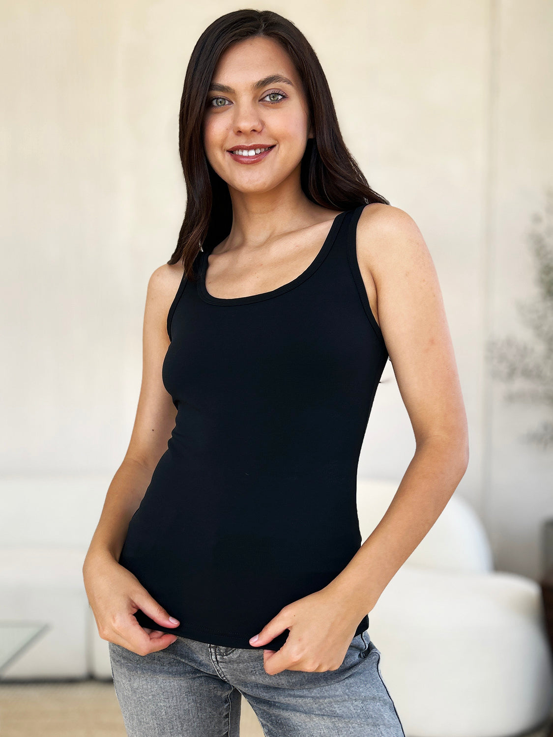 A woman with brown hair, wearing sunglasses, blue jeans, and the Basic Bae Full Size Square Neck Wide Strap Tank in black stands against a concrete wall, showcasing a basic style.