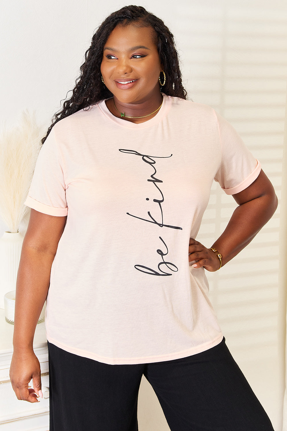 A woman wearing a light pink Simply Love BE KIND Graphic Round Neck T-Shirt with the phrase "be kind" written in cursive. She is standing indoors with a neutral expression, the round neck adding a touch of casual elegance.