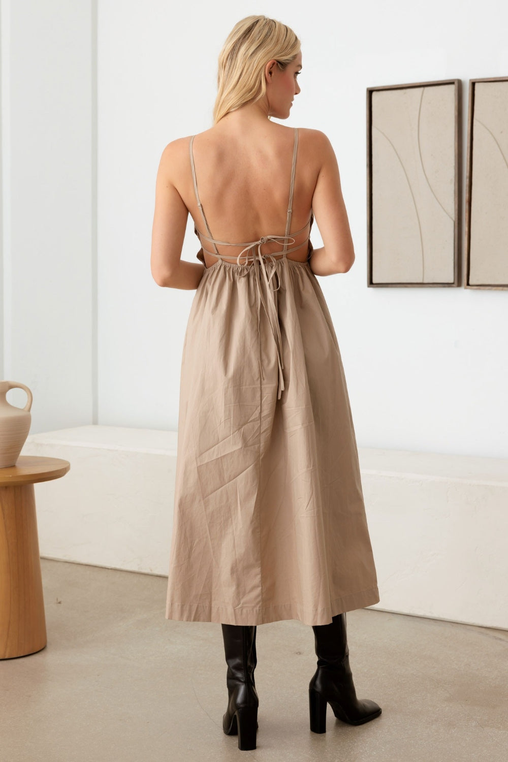 A woman with long blonde hair wears a beige Le Lis Tie Back Backless Cami Dress made from 100% cotton and black knee-high boots. She stands indoors next to a wooden stool with a ceramic vase, while simple wall art adorns the background.