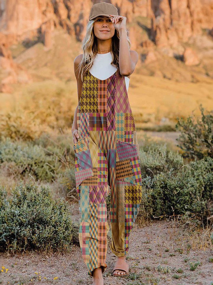 A woman stands outdoors in front of a rocky landscape, wearing the Full Size Printed V-Neck Sleeveless Jumpsuit and a brown brimmed hat. She is smiling and holding the brim of her hat with one hand, exuding the carefree vibe of this vibrant and colorful jumpsuit.