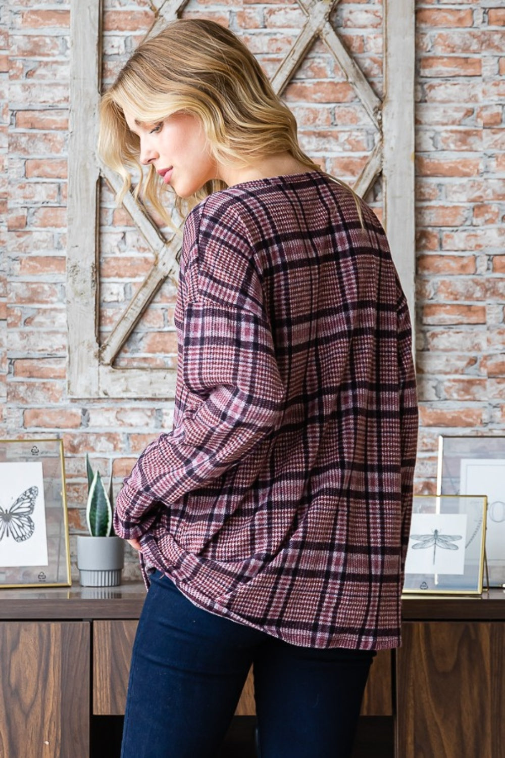 Sporting long blonde hair, a person stands against a brick wall wearing the Heimish Full Size Plaid V-Neck Long Sleeve Top in pink and black. Framed butterfly prints adorn the background, while this adaptable wardrobe item adds a touch of flair to any ensemble.