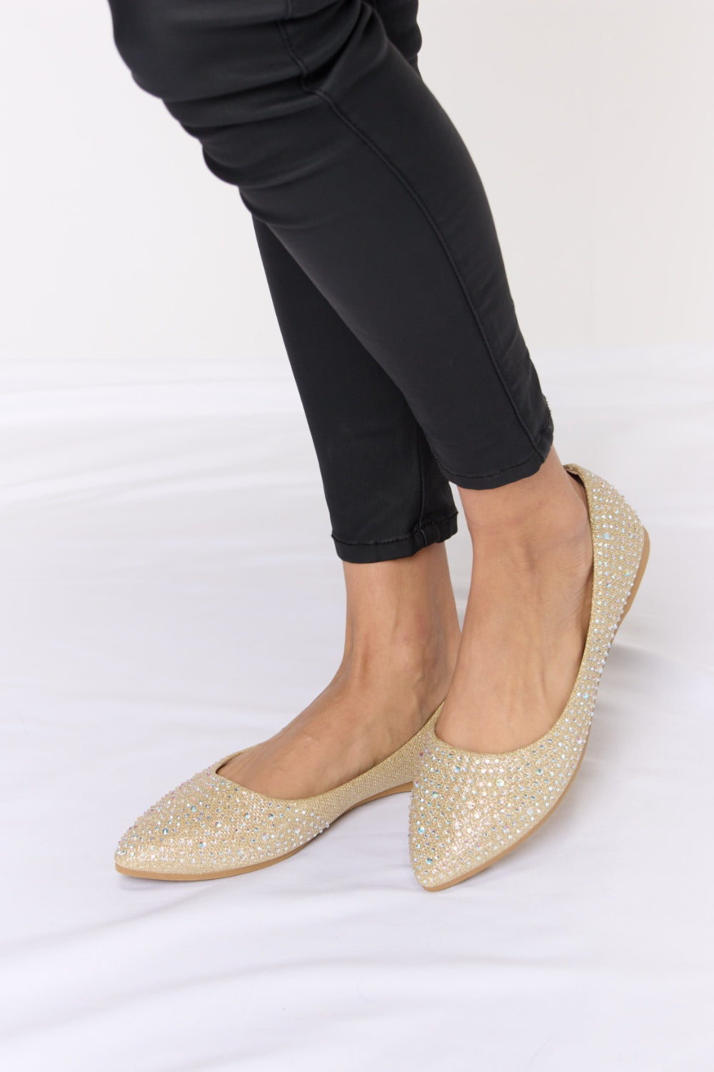 Someone is sitting on a white surface wearing black pants and chic Forever Link Rhinestone Point Toe Flat Slip-Ons in sparkling gold.