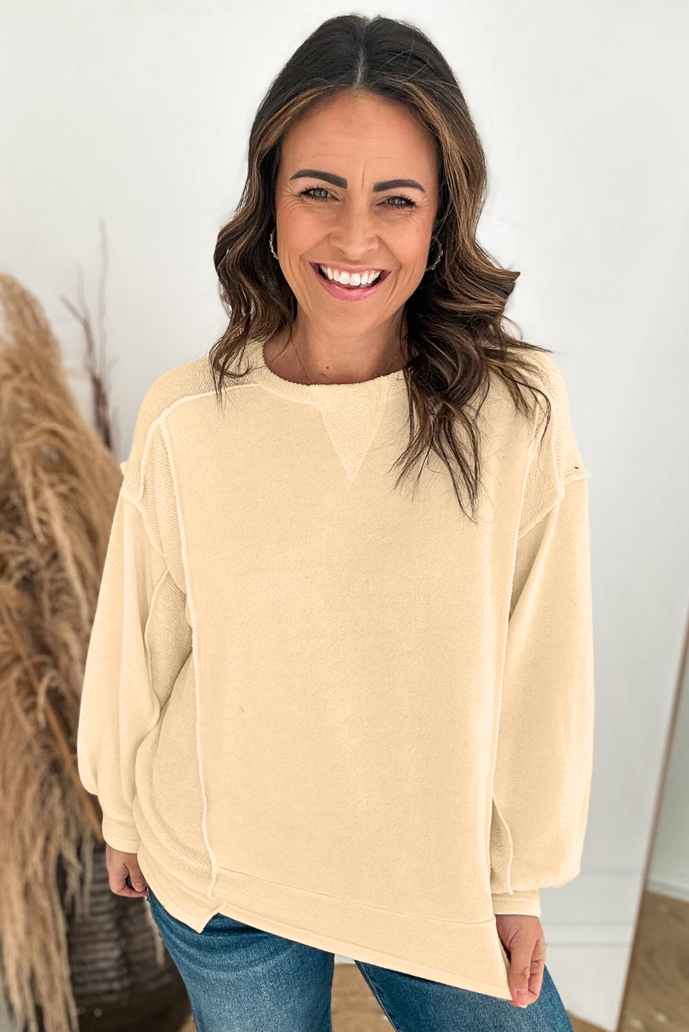 A woman with wavy hair stands with her back to the camera, wearing a Beige Splicing Round Neck Pullover Sweatshirt and blue jeans. She is in front of a white wall and a decorative plant, presenting a casual yet polished look.