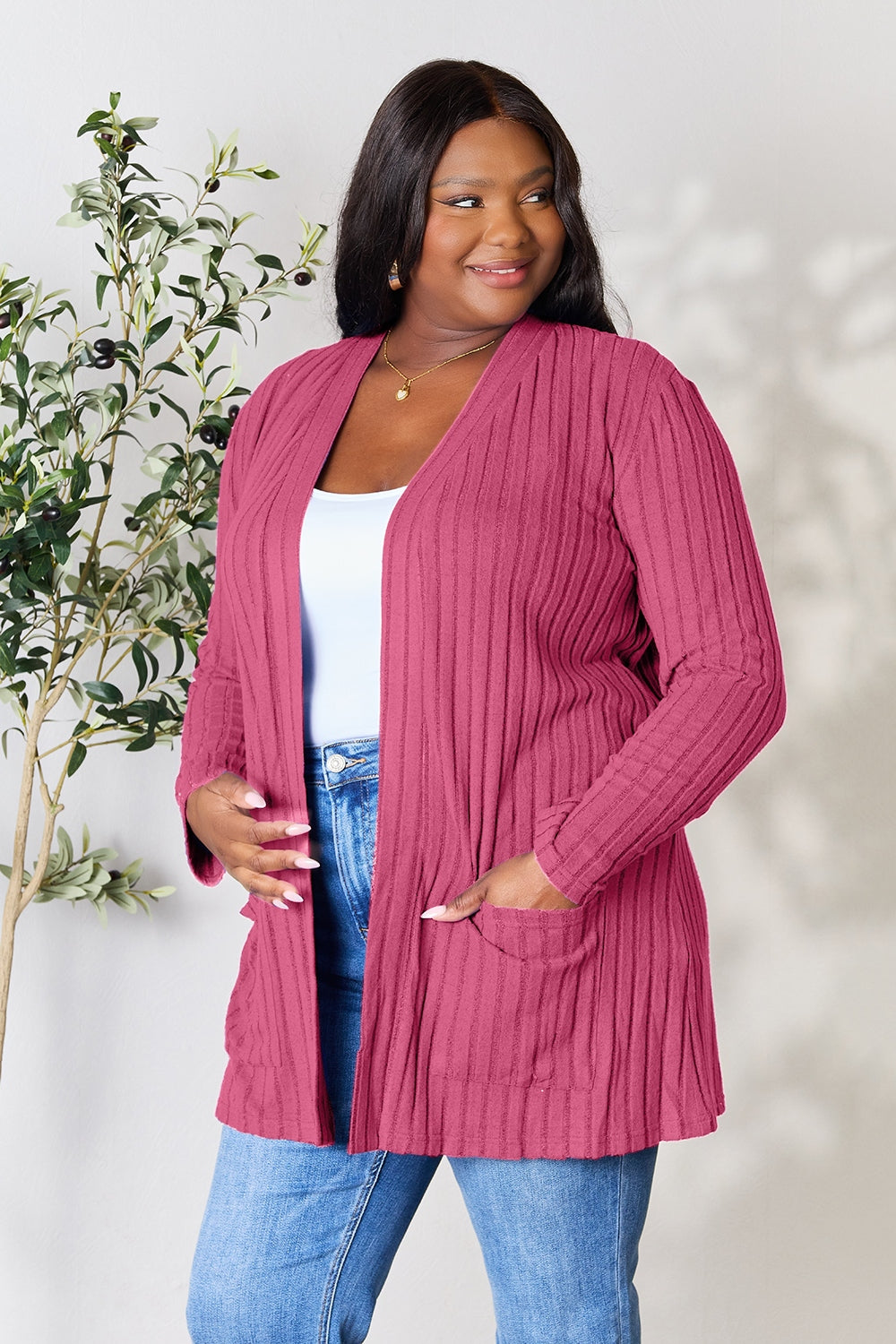 A woman in a Basic Bae Full Size Ribbed Open Front Cardigan with Pockets and jeans, made from stretchy material, stands in front of a plant, smiling slightly and looking to the side.