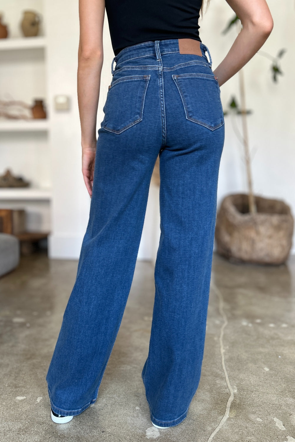 A person wearing Judy Blue Full Size High Rise Straight Jeans, a black top, and white shoes stands gracefully on a polished concrete floor.