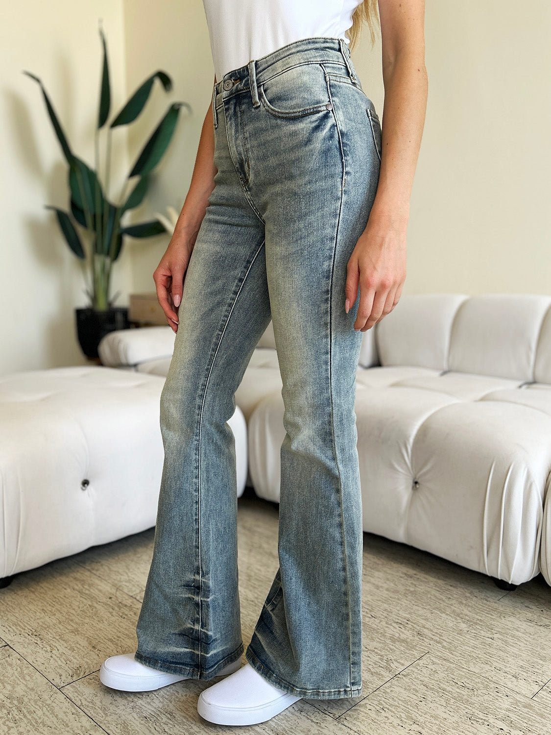 A person wearing Judy Blue Full Size Mid Rise Flare Jeans in a retro-inspired light blue color, paired with a white top and white shoes, stands in a modern living room with a white tufted sofa in the background.