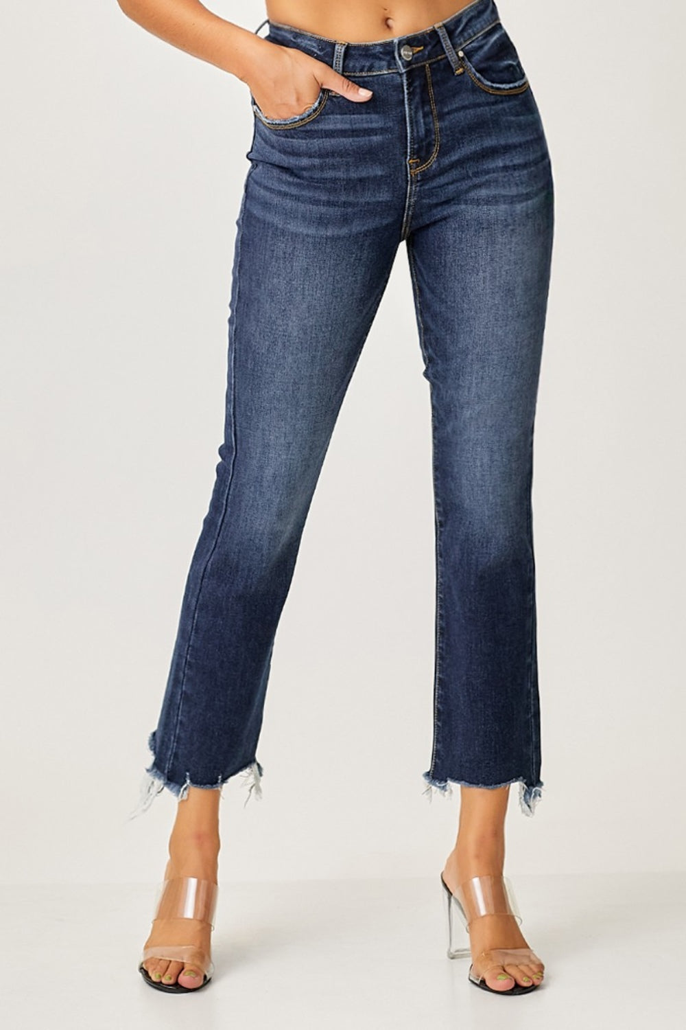 A person striking a pose in Risen Full Size Frayed Hem Cropped Straight Jeans with clear high-heeled sandals, one hand casually tucked in a pocket against a plain background.