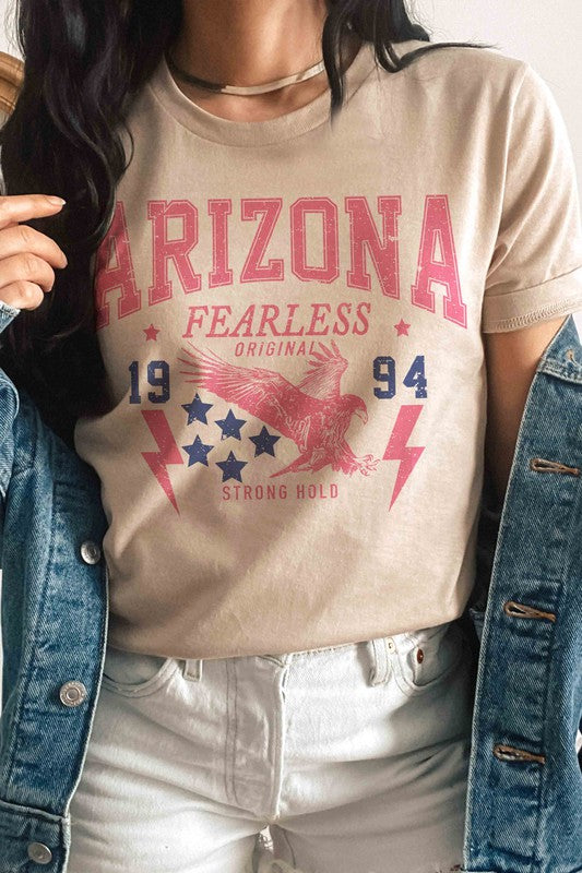 A woman with long light hair is wearing jeans and a 100% cotton ARIZONA EAGLE 1994 Graphic Tee, which features the text "ARIZONA FEARLESS ORIGINAL 1994 STRONG HOLD" along with an eagle, stars, and lightning bolts.