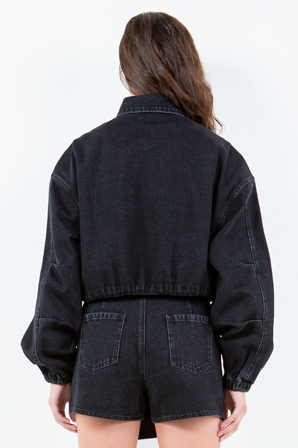 A person with wavy hair is wearing an American Bazi Drawstring Waist Crop Denim Jacket in black, featuring a zippered front closure, paired with a matching black denim skirt.