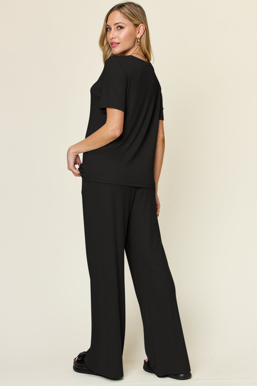 A person stands wearing the Double Take Full Size Round Neck Short Sleeve T-Shirt and Wide Leg Pants Set in a stretchy light grey fabric, with one hand in a pocket and black sandals.