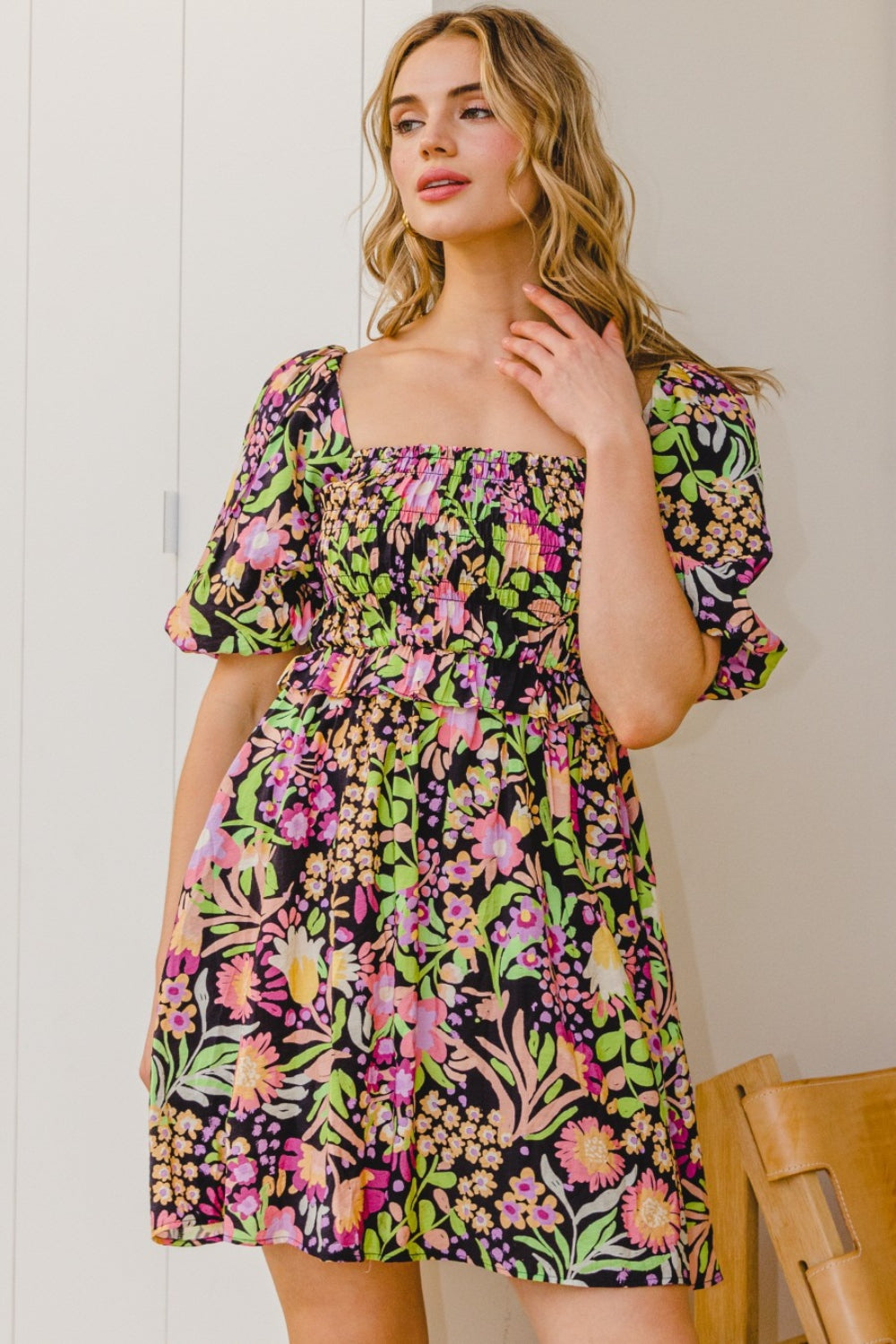 A person stands wearing the ODDI Full Size Floral Tie-Back Mini Dress, a feminine and flirty black floral design with puffy sleeves, perfect for a summer wedding.