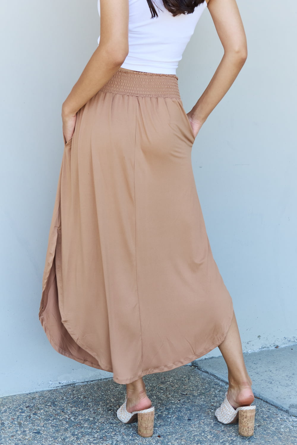 A person stands against a wall, wearing a white sleeveless top, the Doublju Comfort Princess Full Size High Waist Scoop Hem Maxi Skirt in Tan, and blue platform sandals. The right hand rests on the side of the skirt.