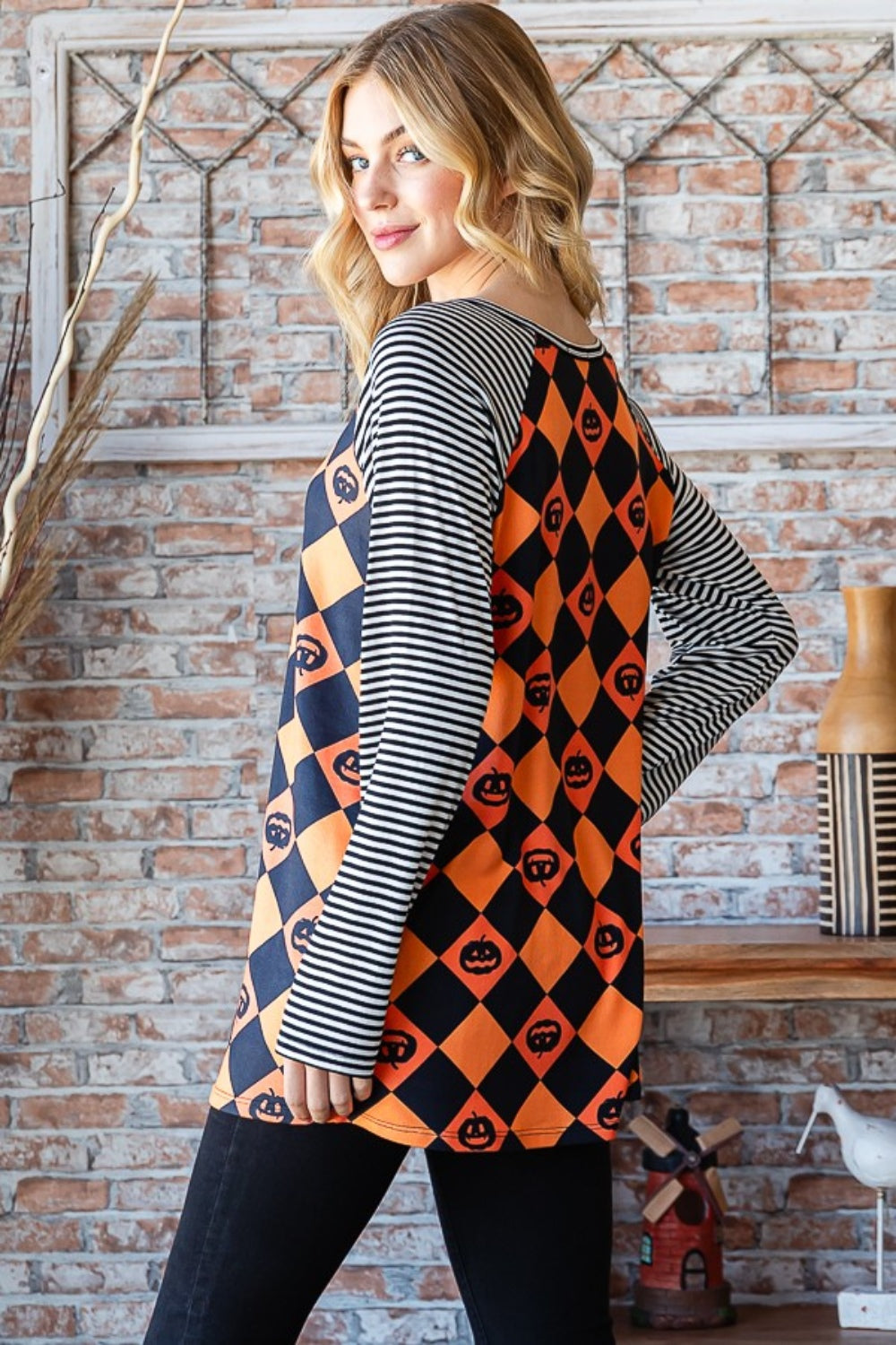 A person wearing the Heimish Full Size Checkered Contrast Striped Long Sleeve T-Shirt, which boasts a casual style with a black and orange checkered pattern adorned with pumpkin designs and contrasting black and white striped sleeves, stands before a brick wall enhanced by dried branches in the backdrop.