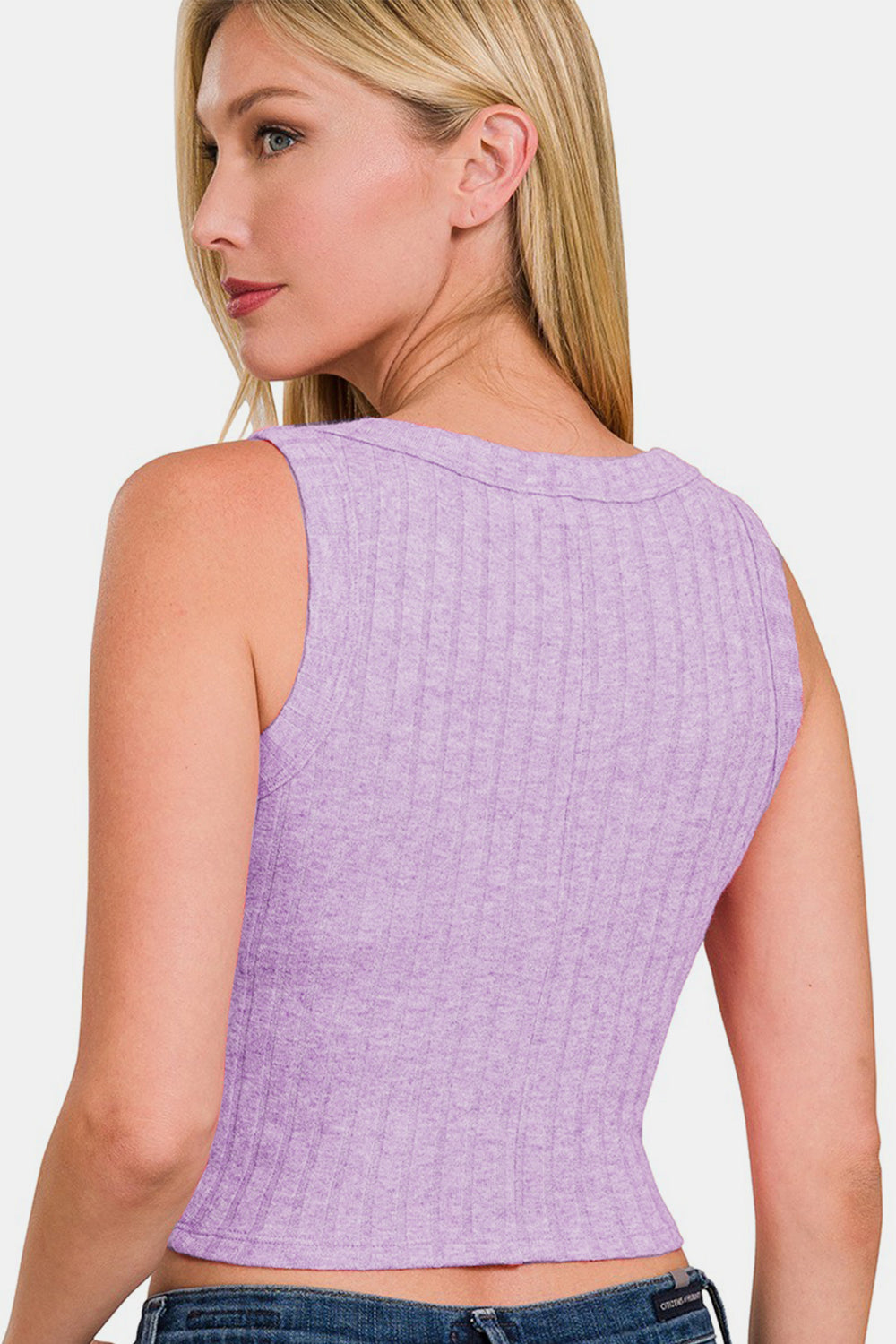 A woman with long blonde hair is wearing a stylish Zenana Ribbed Cropped Tank in lilac paired with jeans against a plain background. She strikes a fashionable pose, placing one hand near her head, effortlessly showcasing her versatile wardrobe.