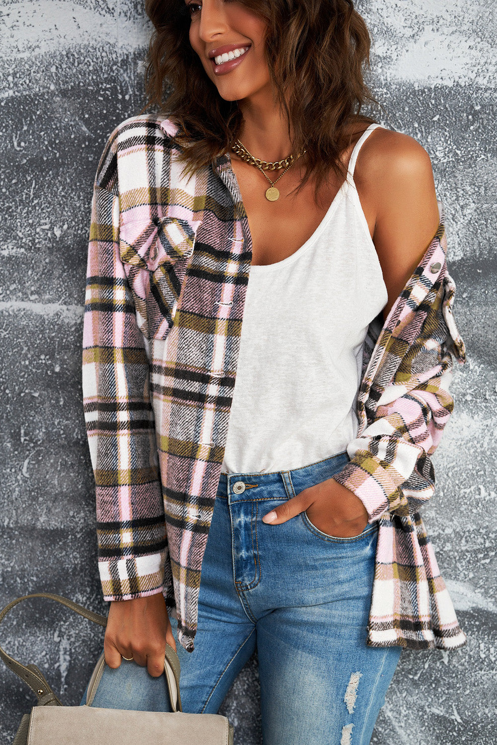 A person wearing a Brown Plaid Button Up Long Sleeve Flannel Shacket layered over a white top with ripped jeans and holding a brown handbag stands against a textured gray wall.