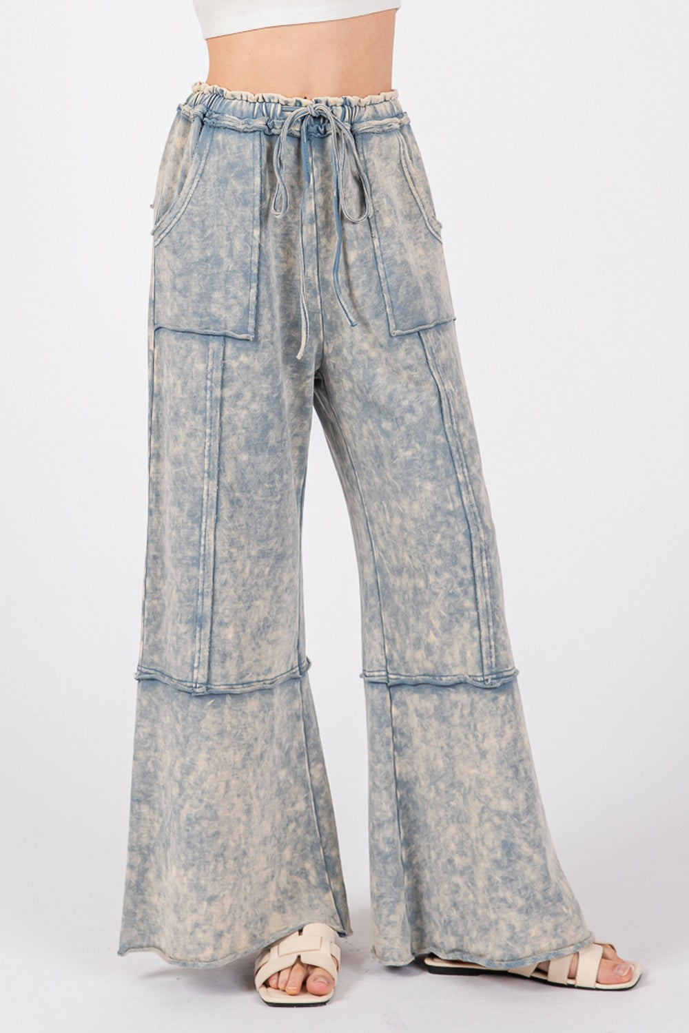 A person is wearing the SAGE + FIG Mineral Washed Terry Wide Leg Pants, which are high-waisted and light blue with a tie-dye appearance. Made from breathable cotton, they feature a drawstring elastic waistband and large front pockets, paired with beige sandals.