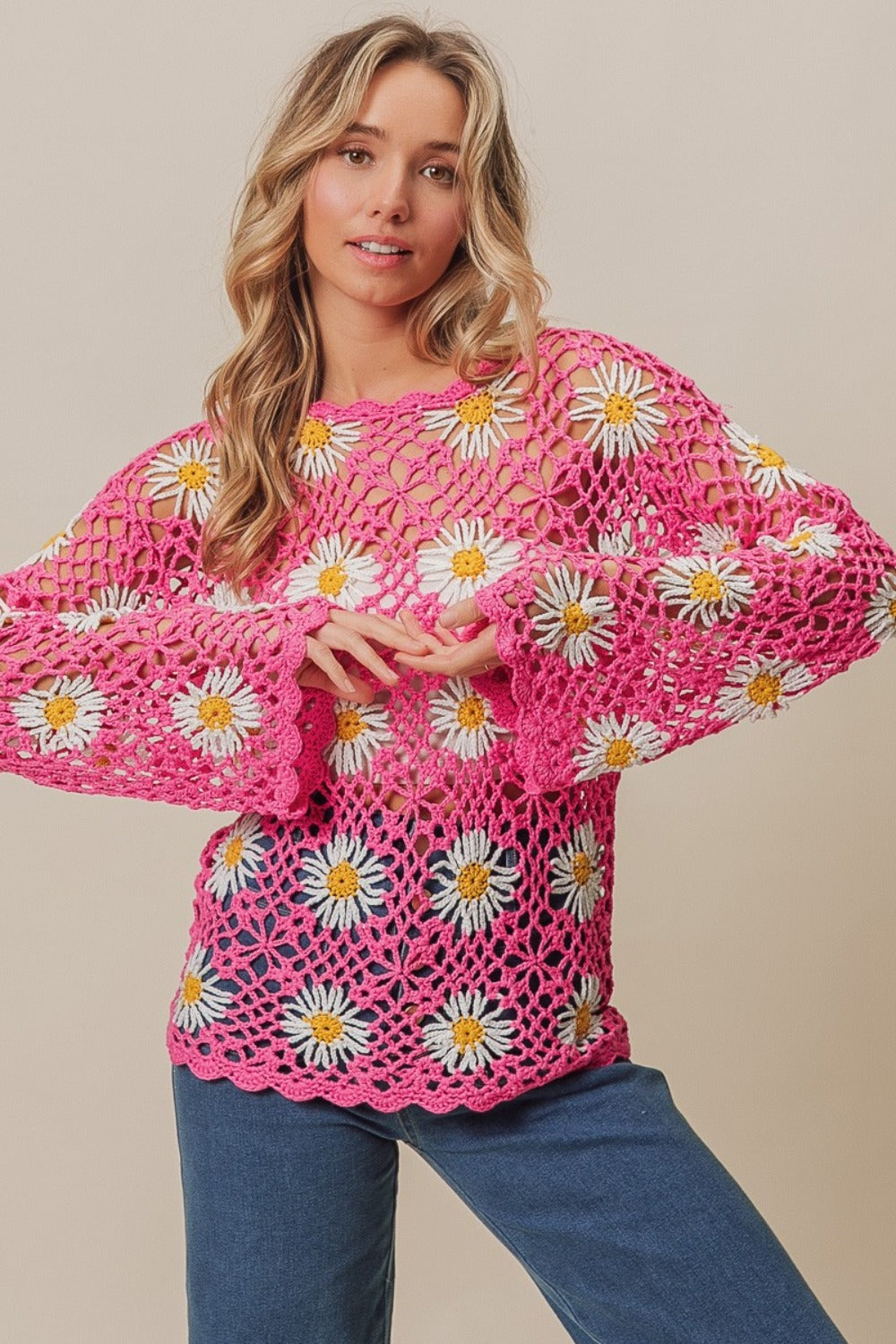 A person stands wearing the BiBi Floral Crochet Net Lace Cover Up, featuring pink crochet work adorned with white and yellow daisy patterns, exuding Bohemian style. They have long, wavy hair and pose with hands together.