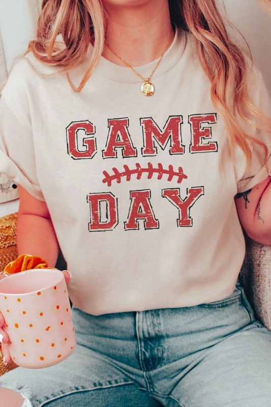 A young woman with long brown hair is wearing a 100% cotton GAME DAY Graphic Tee, featuring a football graphic and "Game Day" text, as she stands against a neutral background.