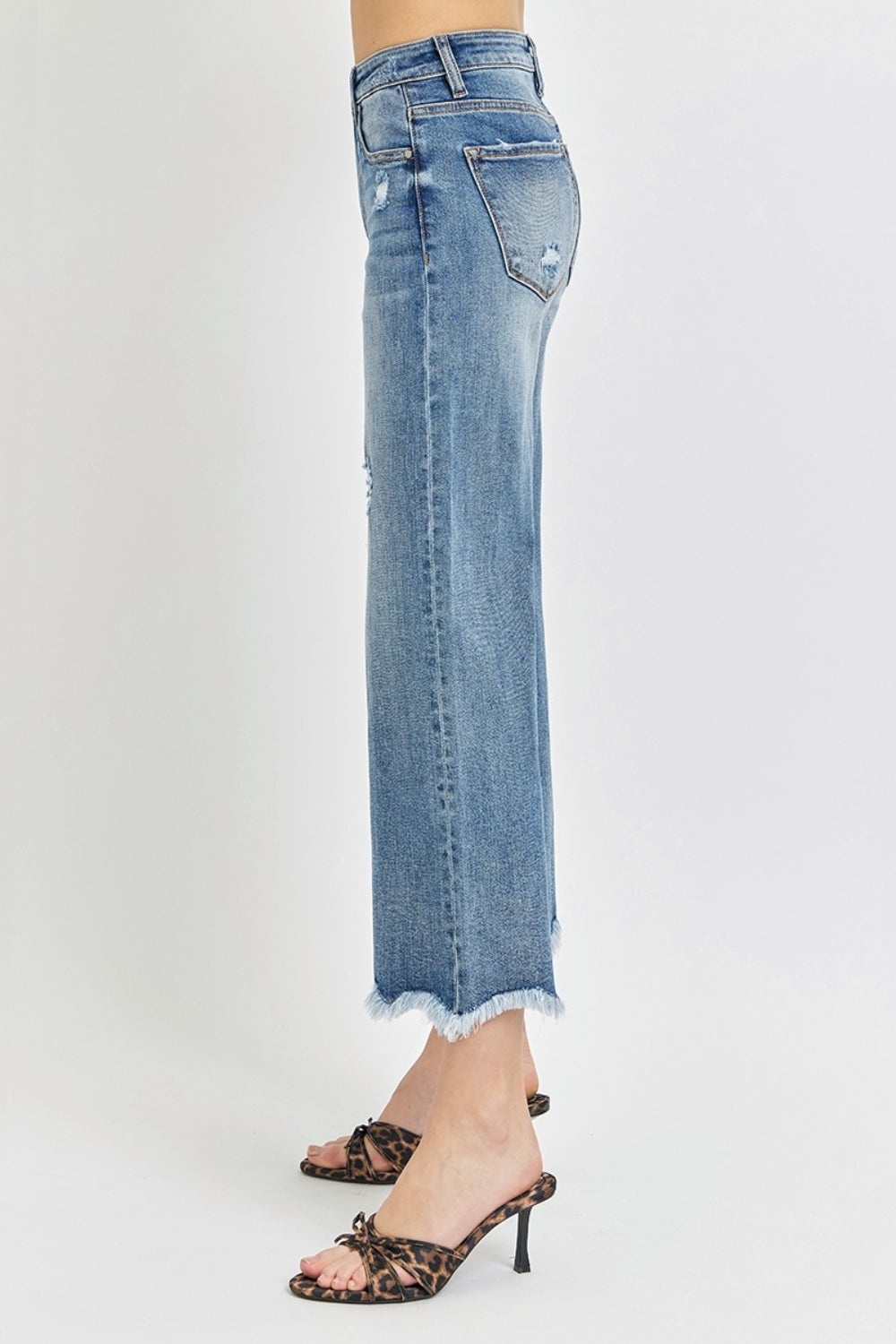 Person wearing Risen High Rise Cropped Flare Jeans with frayed details and a few rips, paired with strappy high-heeled sandals against a plain background.