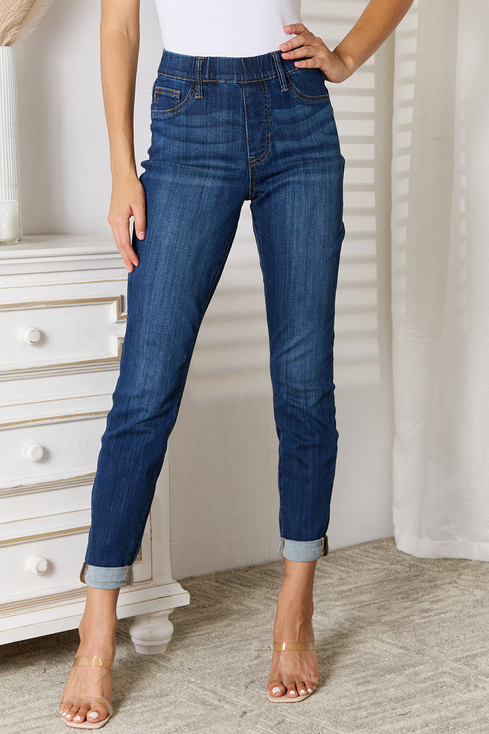 A person wearing Judy Blue Full Size Skinny Cropped Jeans with rolled-up cuffs and clear heels stands near a white dresser adorned with a decorative vase for a modern look.