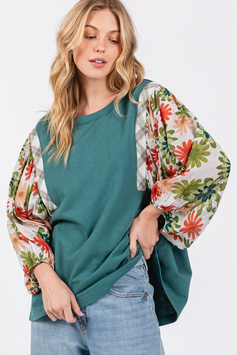 A person in a hat wears the SAGE + FIG Full Size Printed Balloon Sleeve Contrast Top with teal floral-patterned sleeves, blue jeans, and a back heart patch detail.