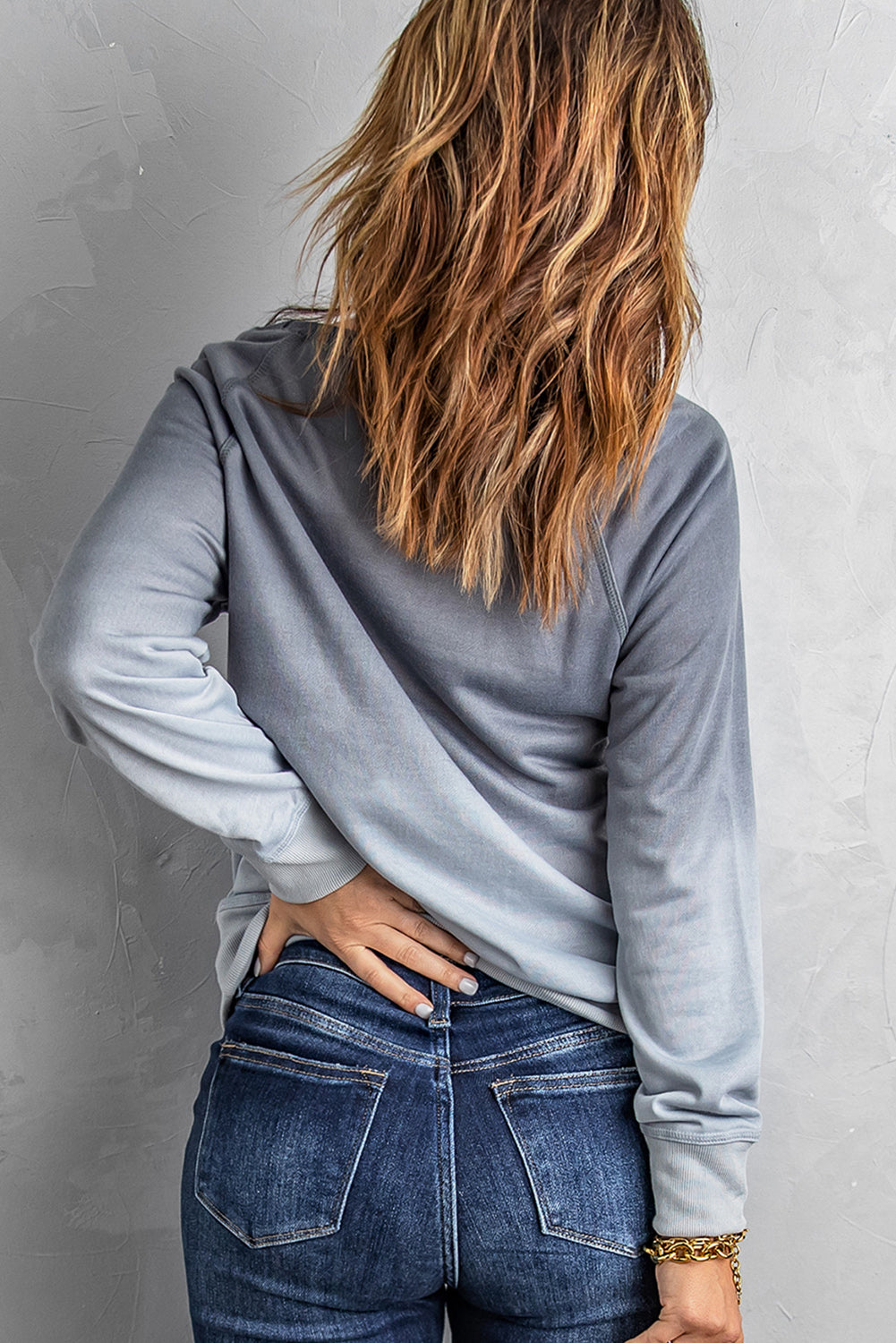 A person with wavy hair stands facing a gray wall, wearing a Gray Ombre Crewneck Long Sleeve Sweatshirt and blue jeans, with one hand on their hip.