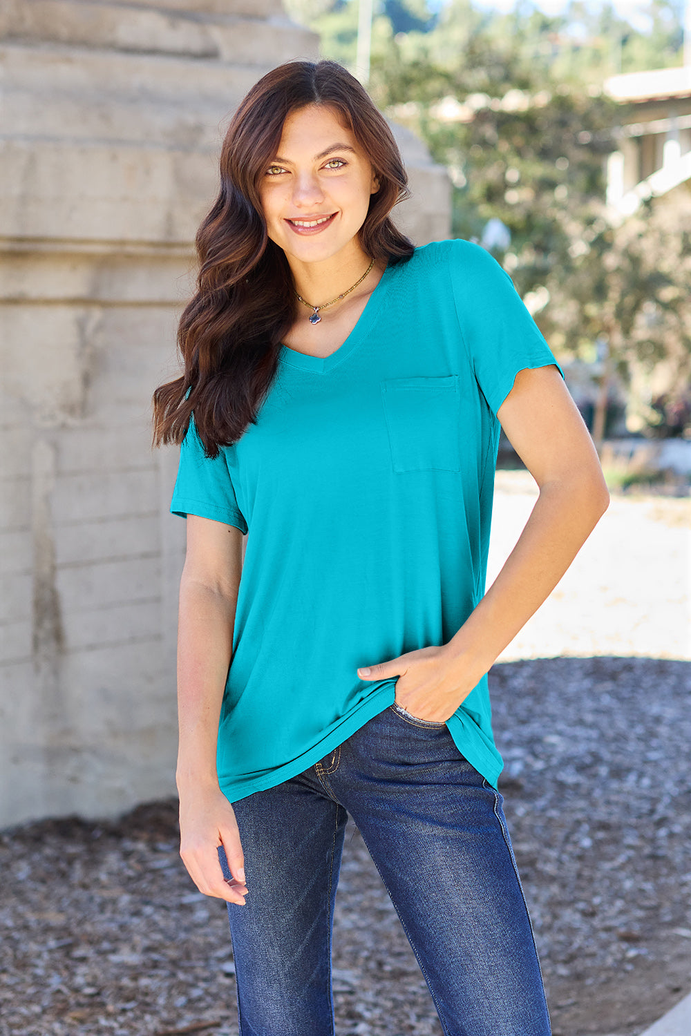 A woman with long brown hair, wearing a Basic Bae Full Size V-Neck Short Sleeve T-Shirt in teal and stretchy blue jeans, stands outdoors near a stone structure, smiling at the camera with one hand in her pocket.