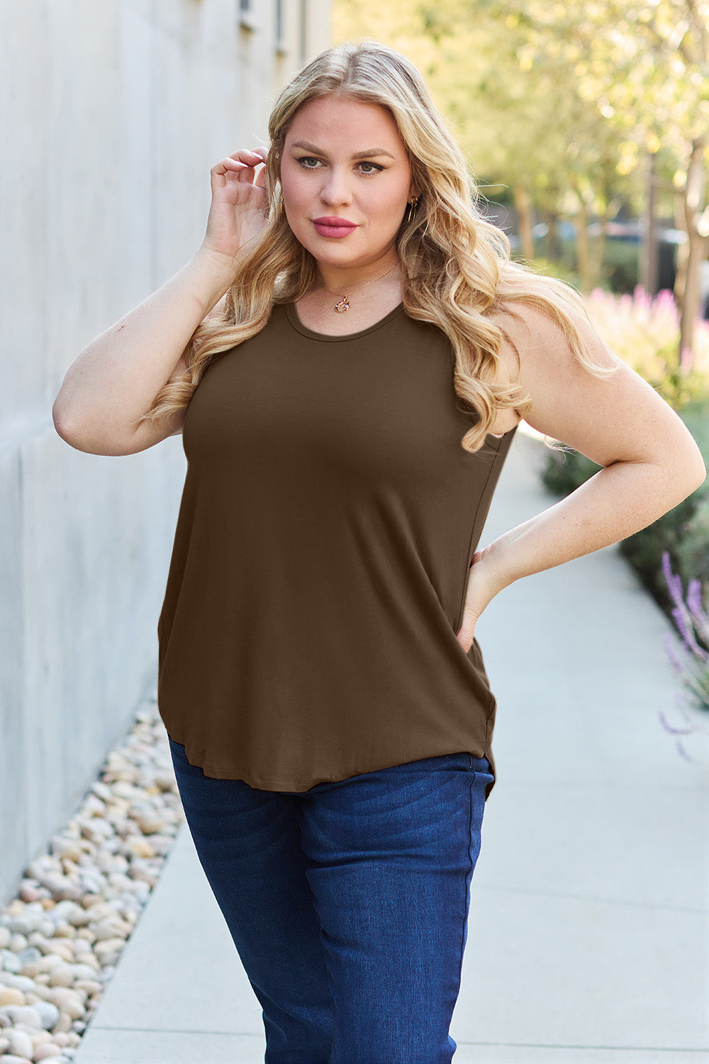 A woman wearing sunglasses, blue jeans, and the Basic Bae Full Size Round Neck Curved Hem Tank is standing in front of a concrete wall, holding her glasses with one hand and carrying a beige shoulder bag. Her basic style complements the slightly stretchy fabric of her outfit, ensuring comfort while maintaining chic simplicity.