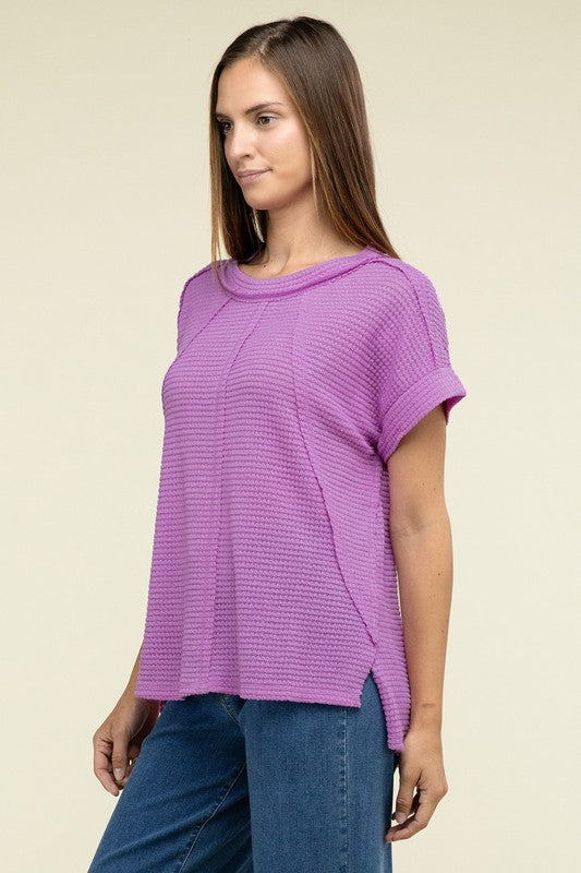 A woman with long brown hair is wearing a purple Brushed Waffle Exposed-Seam Short Sleeve Top and blue jeans, standing against a plain beige background. This casual wardrobe choice, with its short sleeves and side slits, is perfect for everyday wear.