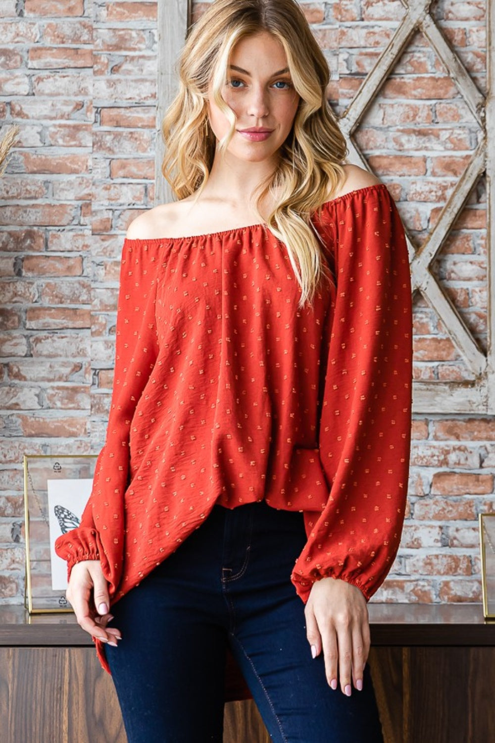 A woman wearing a Heimish Full Size Swiss Dot Off Shoulder Top paired with dark pants poses indoors against a brick wall decorated with butterfly artwork.