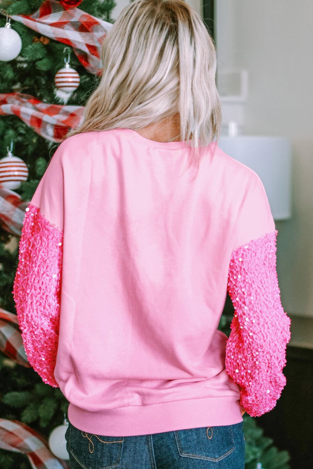 A person with blonde hair wearing a Pink Sequin Patchwork Long Sleeve Pullover Top is standing with their back to the camera near a decorated Christmas tree.