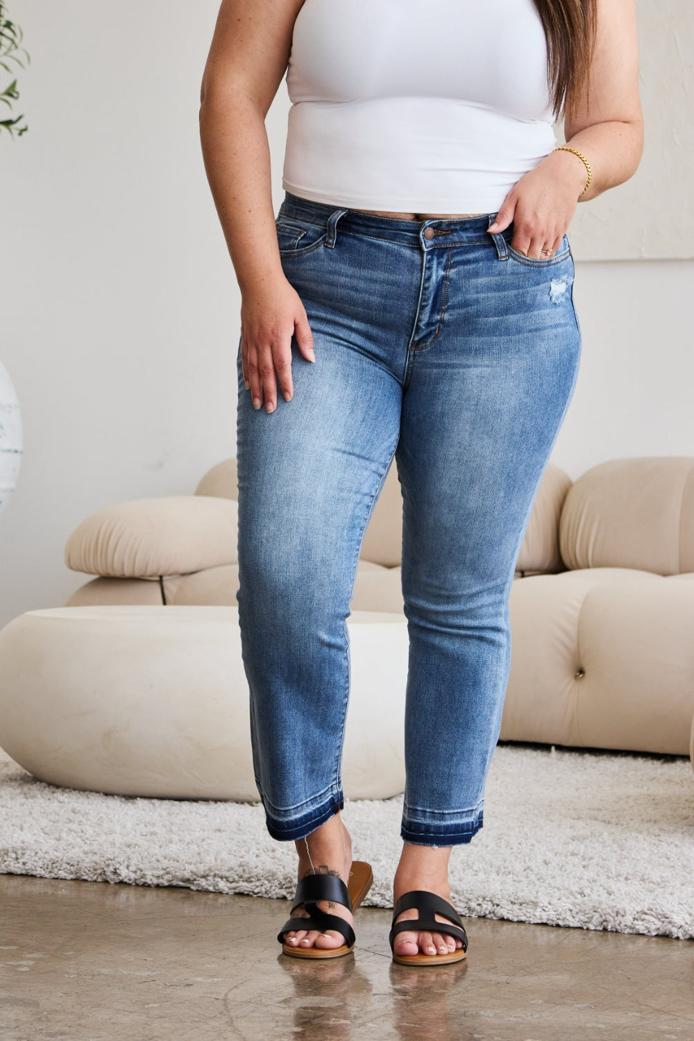 A person modeling Judy Blue Full Size Release Hem Cropped Bootcut Jeans and white heeled sandals stands in a room with light-colored furniture.