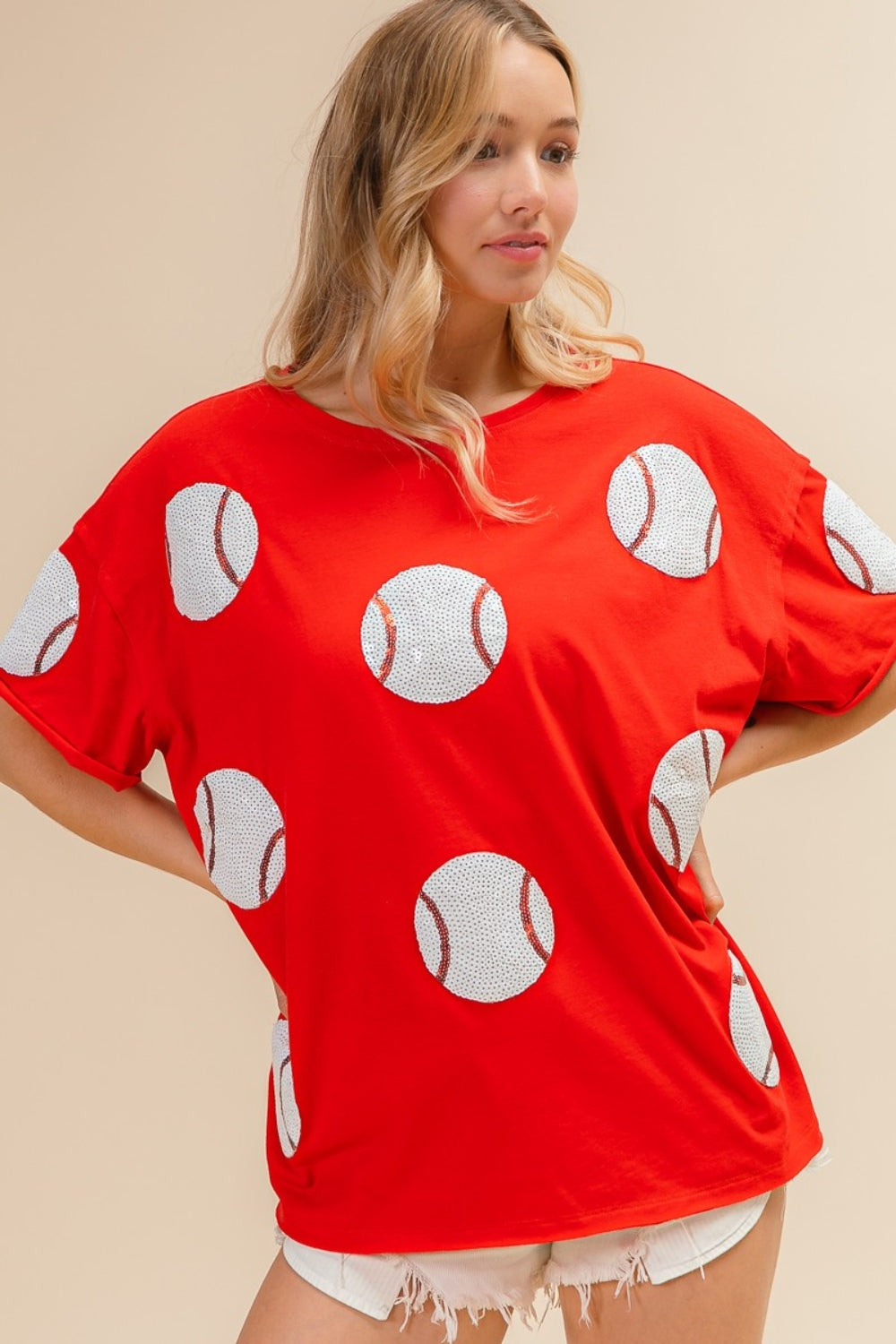 Woman wearing the BiBi Sequin Baseball Drop Shoulder Short Sleeve T-Shirt in red, paired with white shorts, posing against a beige background.